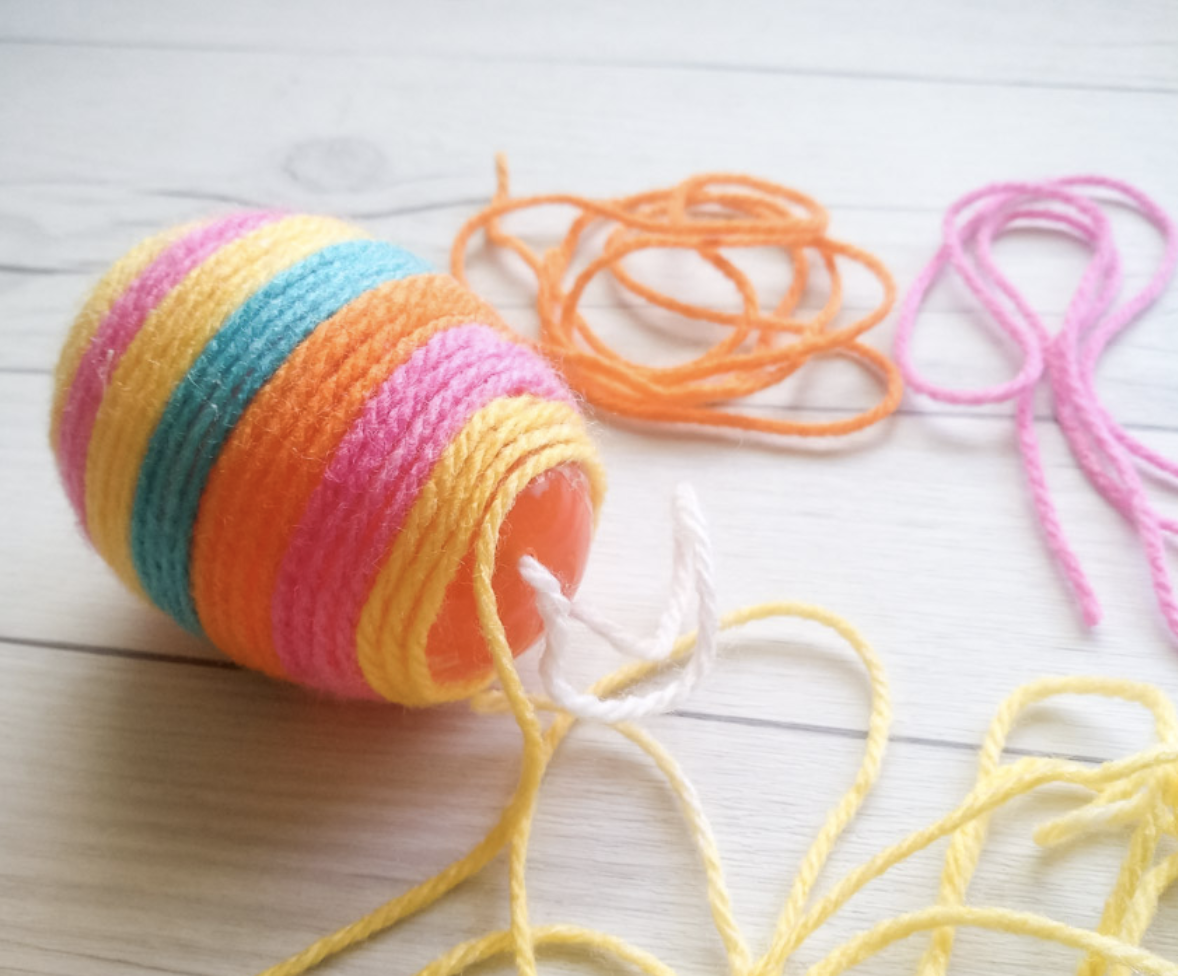 Plastic egg wrapped in colorful yarn with extra strands of yarn scattered on a wooden surface.