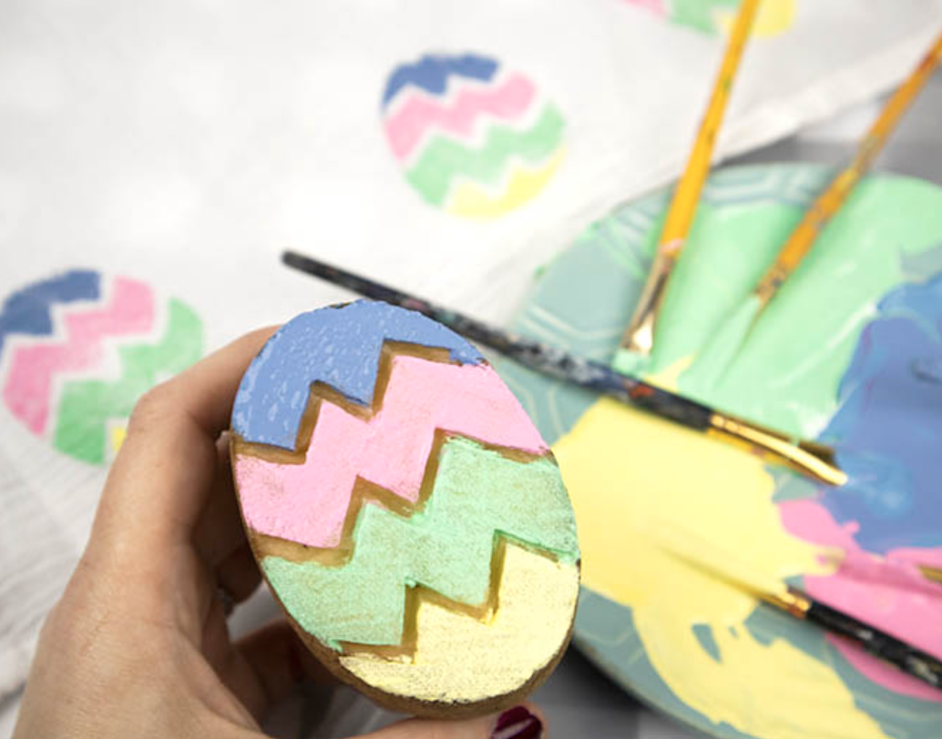 Hand holding a painted Easter egg with pastel zigzag stripes, with paintbrushes and a palette in the background.