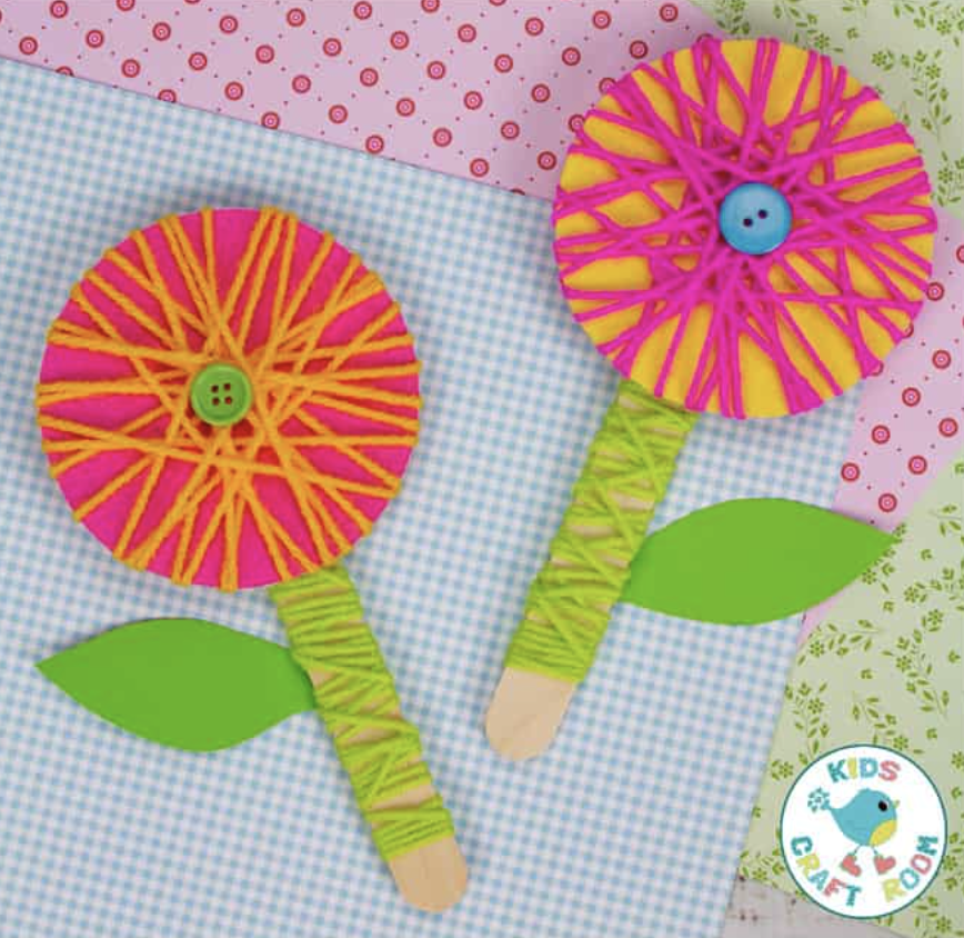 Bright, colorful craft flowers made from yarn-wrapped sticks with paper leaves, displayed on patterned background paper.