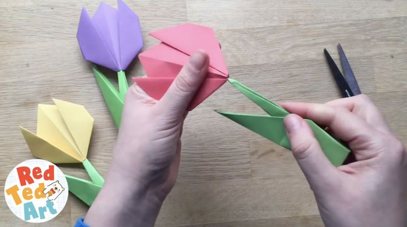 Hands folding a pink origami tulip with green paper stems and scissors nearby.