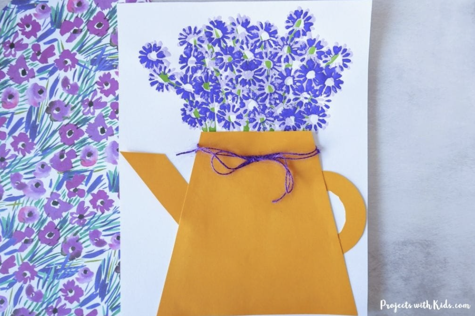 Paper craft of a yellow watering can filled with purple painted flowers, tied with a piece of purple string.