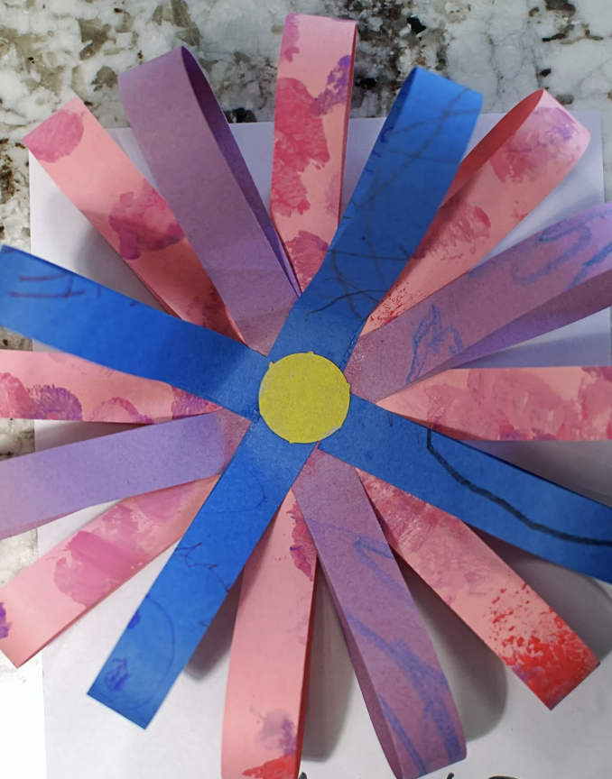 Close-up of a large paper flower made from folded pink, purple, and blue strips with a yellow center.