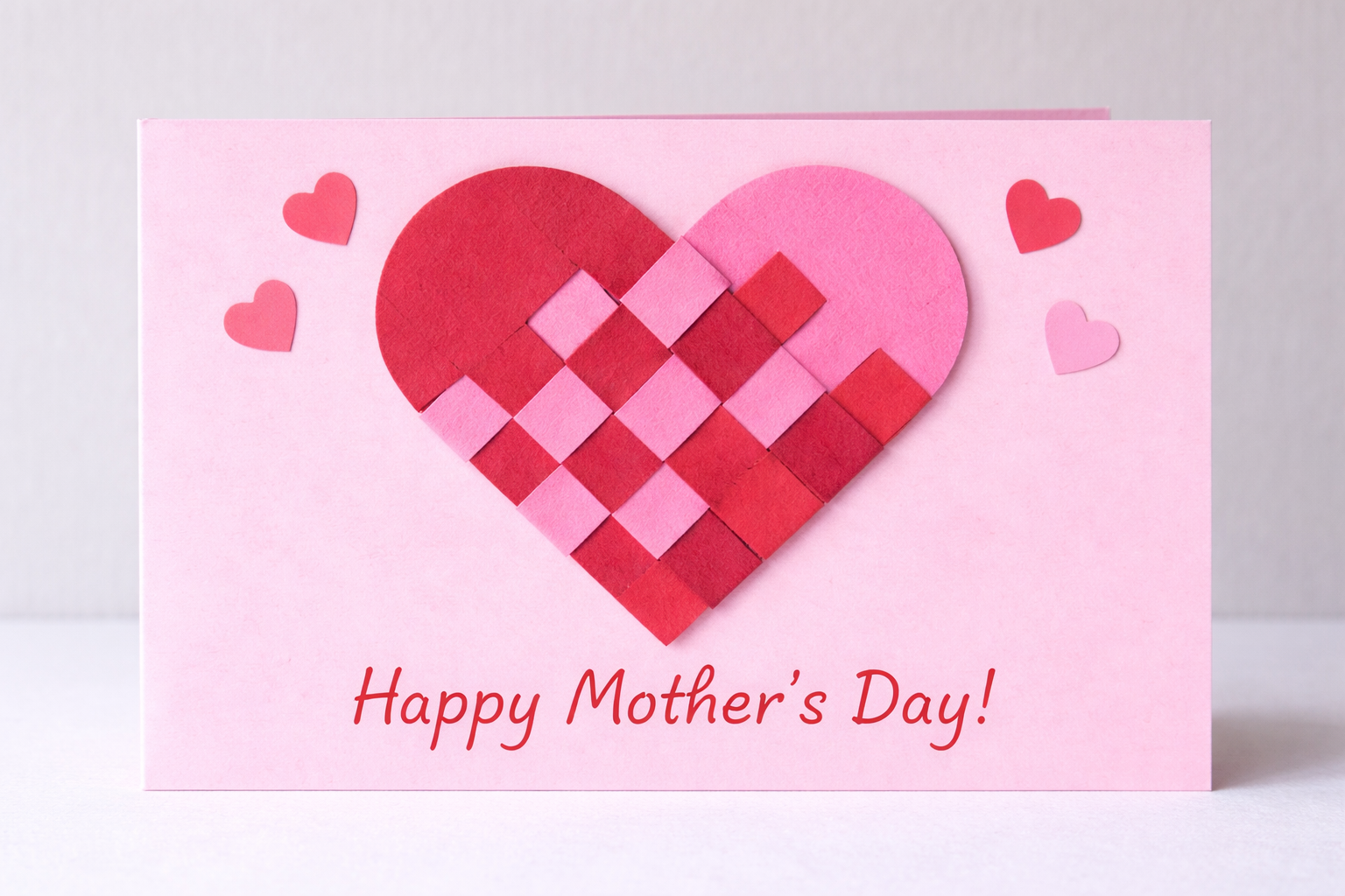 Pink handmade Mother’s Day card featuring a woven paper heart in red and light pink, with small heart decorations and the message “Happy Mother’s Day!” written at the bottom.