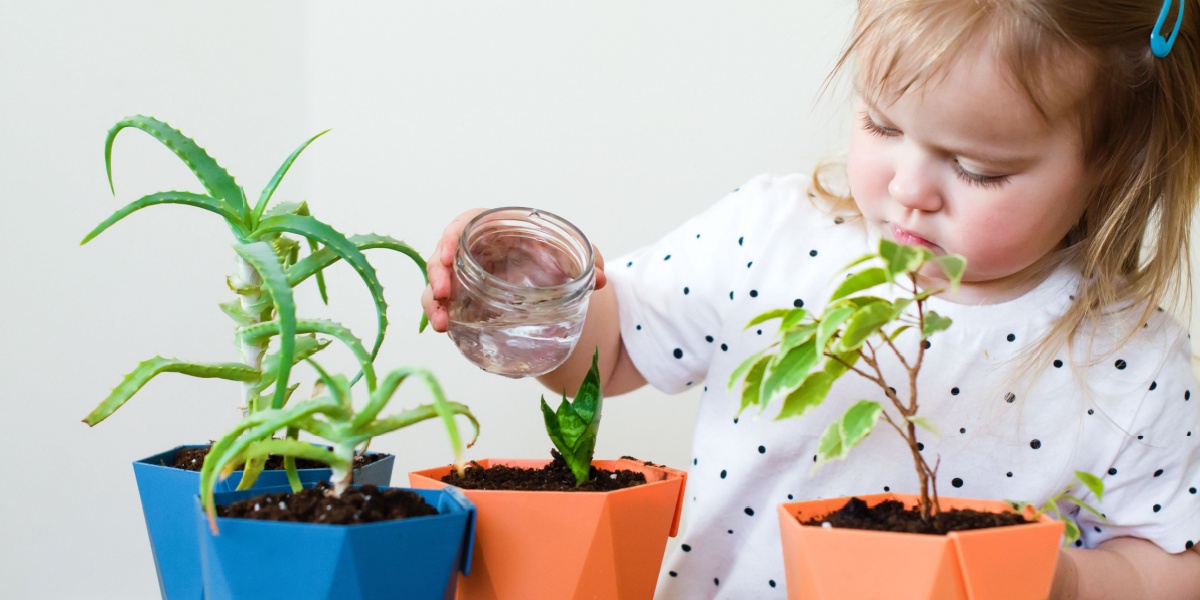 Young child watering small potted plants with a glass jar indoors.