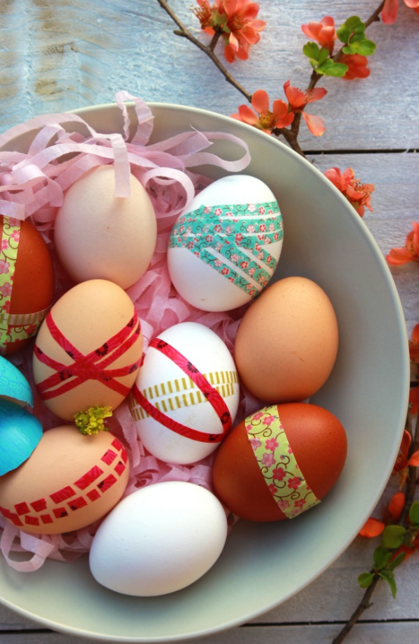 Bowl of decorated Easter eggs featuring colorful washi tape designs, nestled in pink shredded paper with spring flowers nearby.