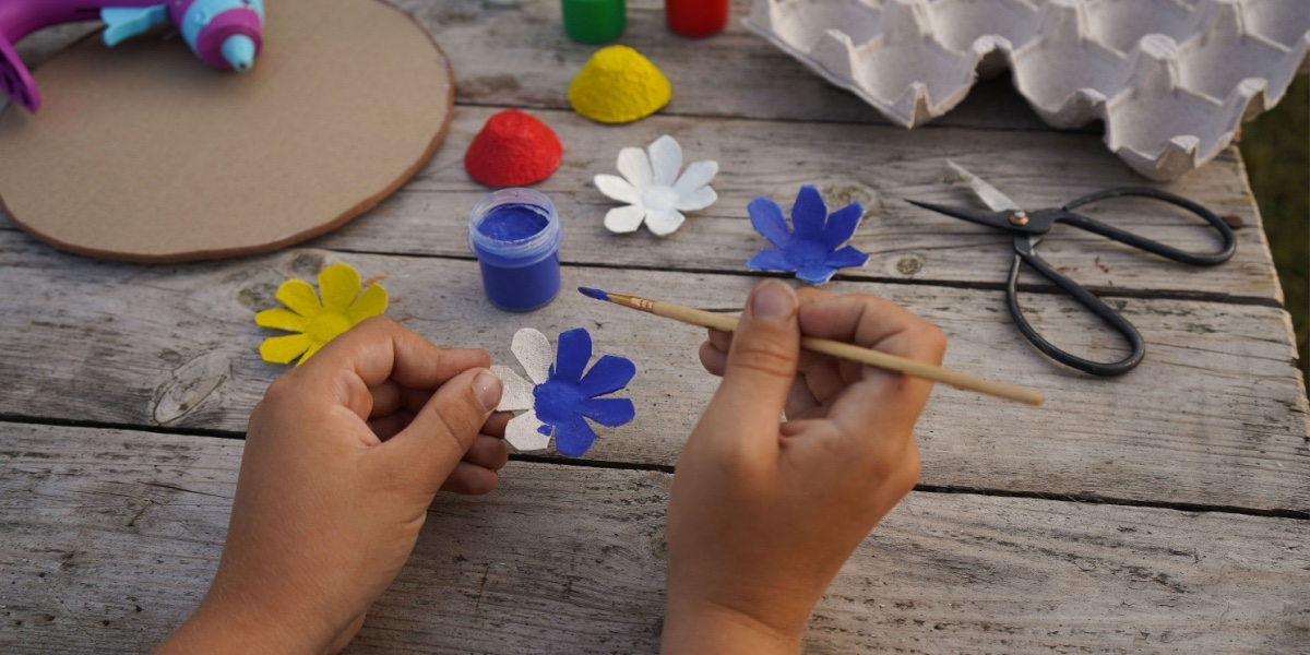 Child painting blue egg carton flower cutouts on a wooden table with paint and craft supplies.