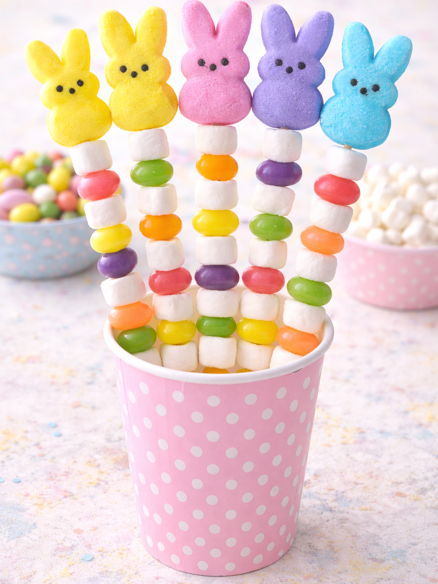 Colorful marshmallow bunny treats stacked on sticks with mini marshmallows and jelly beans, displayed in a pink polka dot cup.