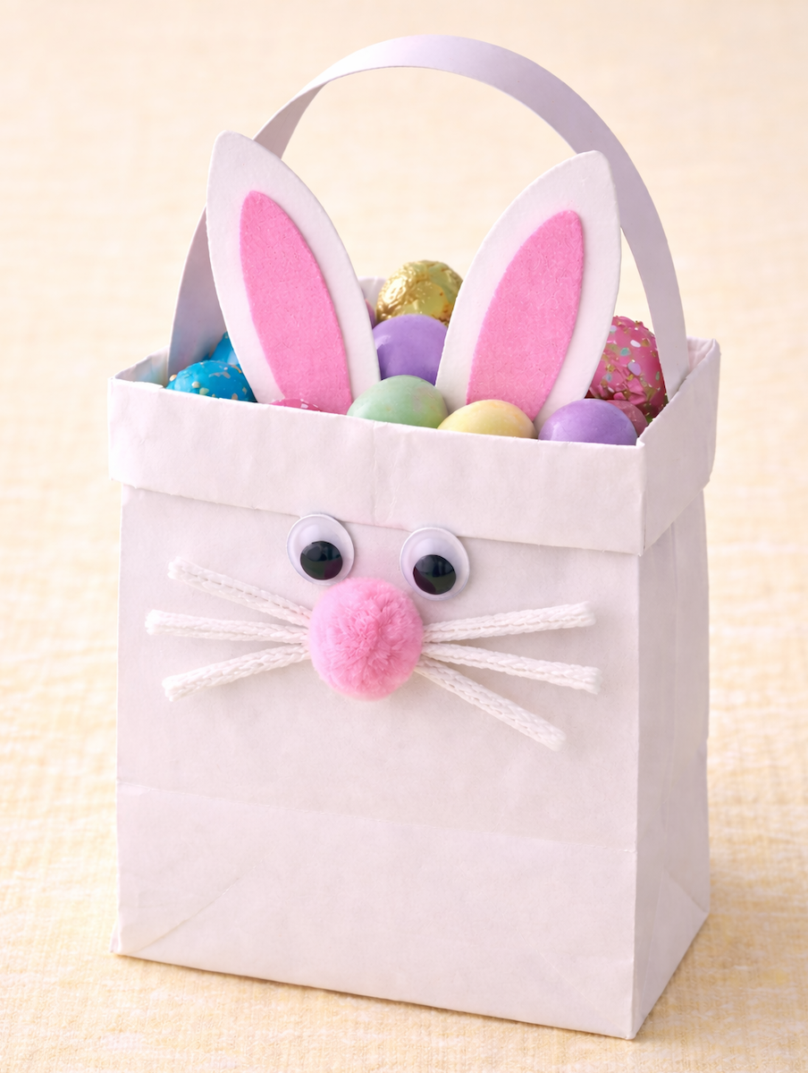 White paper gift bag decorated like a bunny with pink ears, googly eyes, whiskers, and a pom-pom nose, filled with colorful Easter eggs.