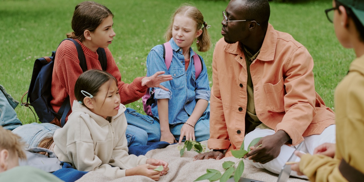 Teacher with a group of four students seated on a blanket outside discussing nature, trees, and different leaves.