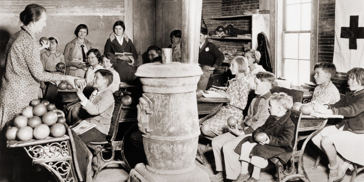 Teacher distributes apples to seated children in a classroom with a large central stove, observing peers and parents in the back room.
