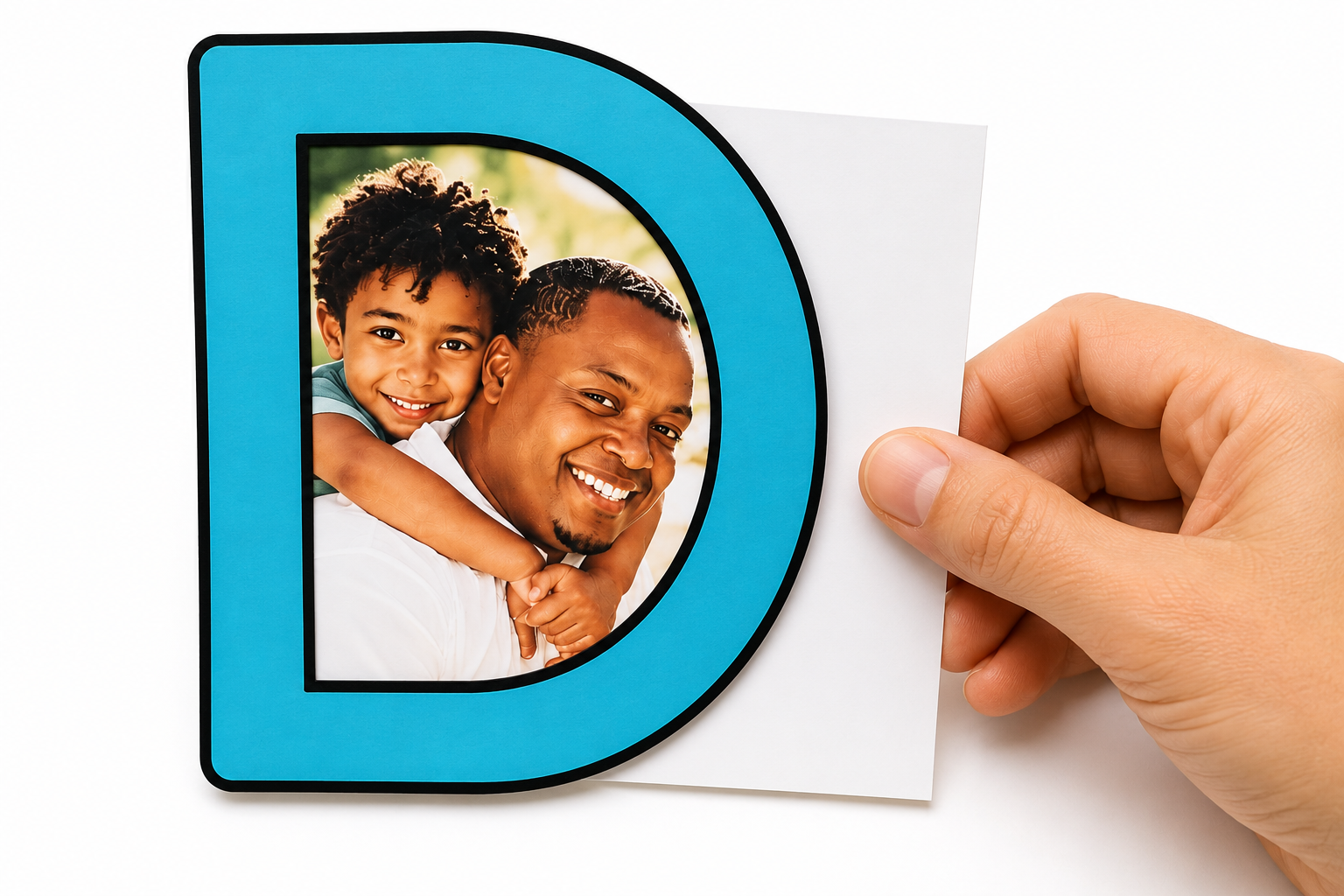 Close-up of a blue letter “D” craft with a father-and-child photo attached behind the cutout, while a hand positions the photo on white paper for a Father’s Day project.