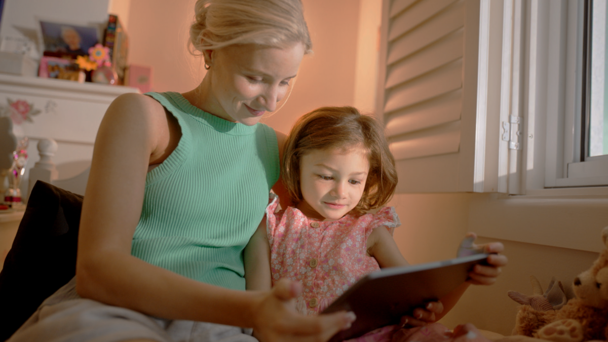 Woman and young girl sit close on a bed, reading a tablet together by a window with warm indoor light.