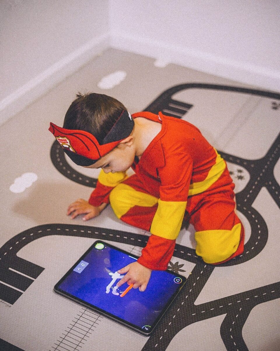 Child in a red and yellow superhero costume kneels on a road-themed play mat, using a tablet with a finger.