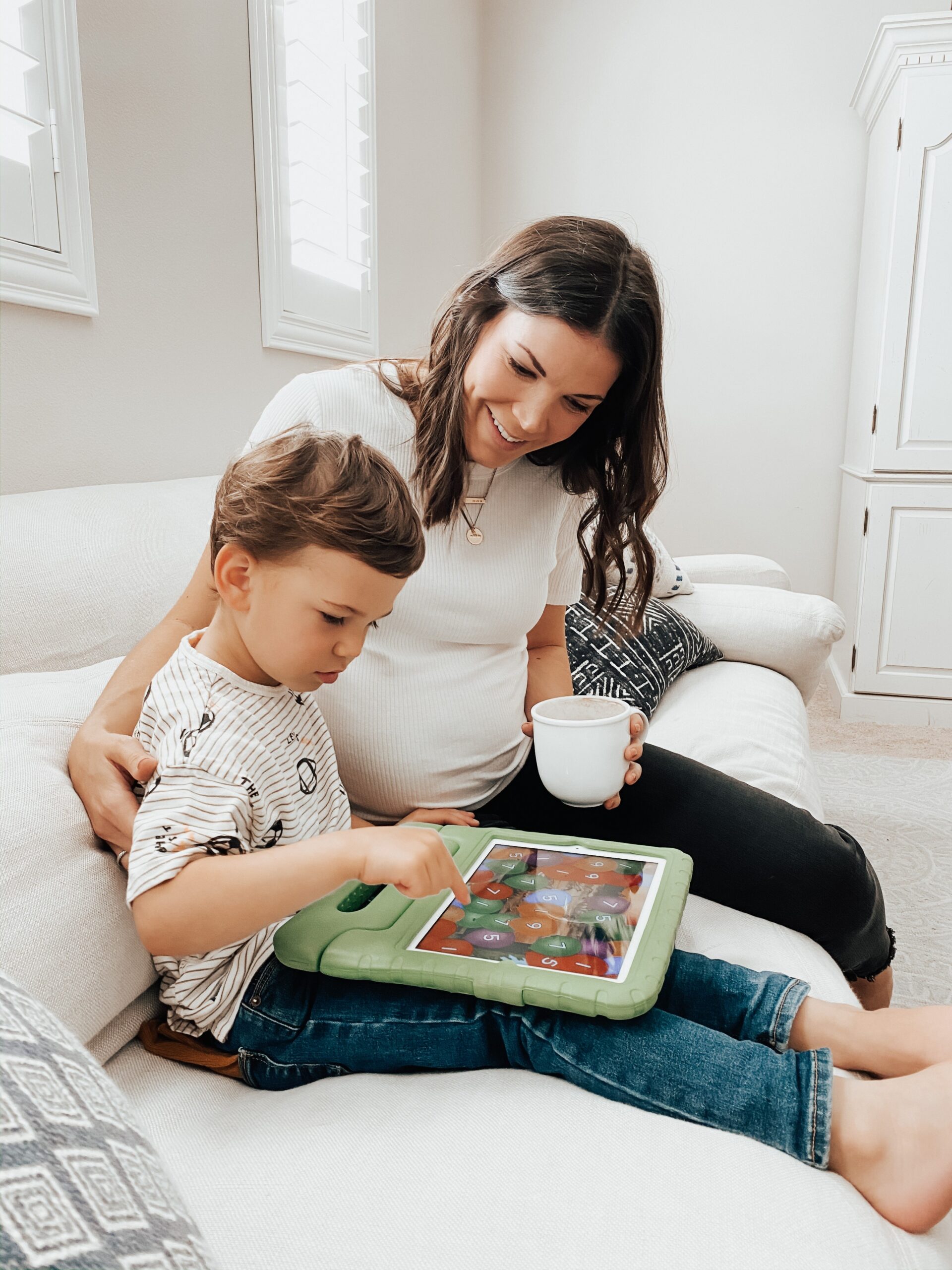 Mother and young boy sit together on a light couch, looking at a tablet screen.