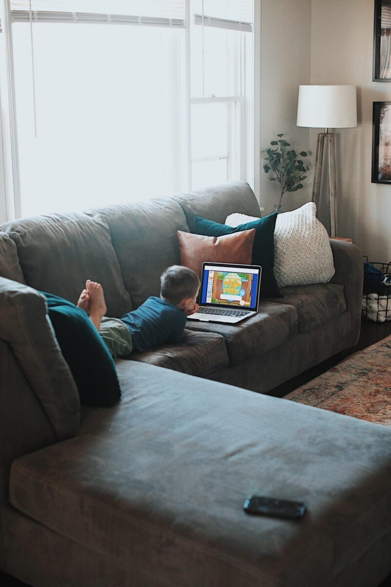 Child lying on a gray sectional sofa using a laptop, with a smartphone nearby, in a bright living room with a lamp and plant nearby.