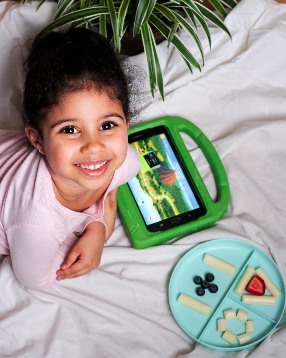 Young girl smiling while lying on white sheets next to a green handheld tablet and a blue divided snack plate with cheese sticks, blueberries, and a slice of watermelon.