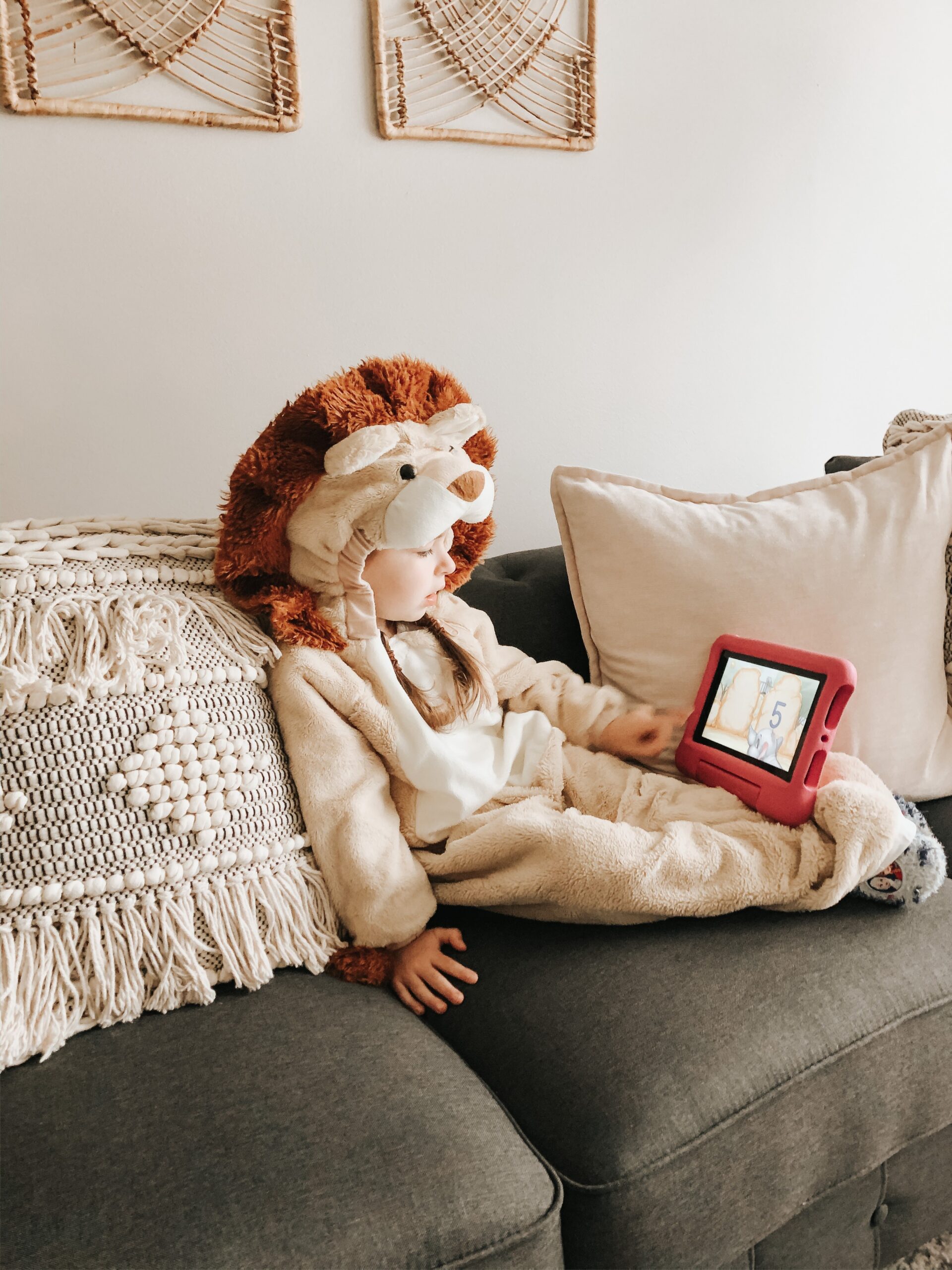 Child in a fluffy lion onesie sitting on a gray sofa, holding a red tablet with a displayed image/game on the screen, cozy beige cushions nearby.