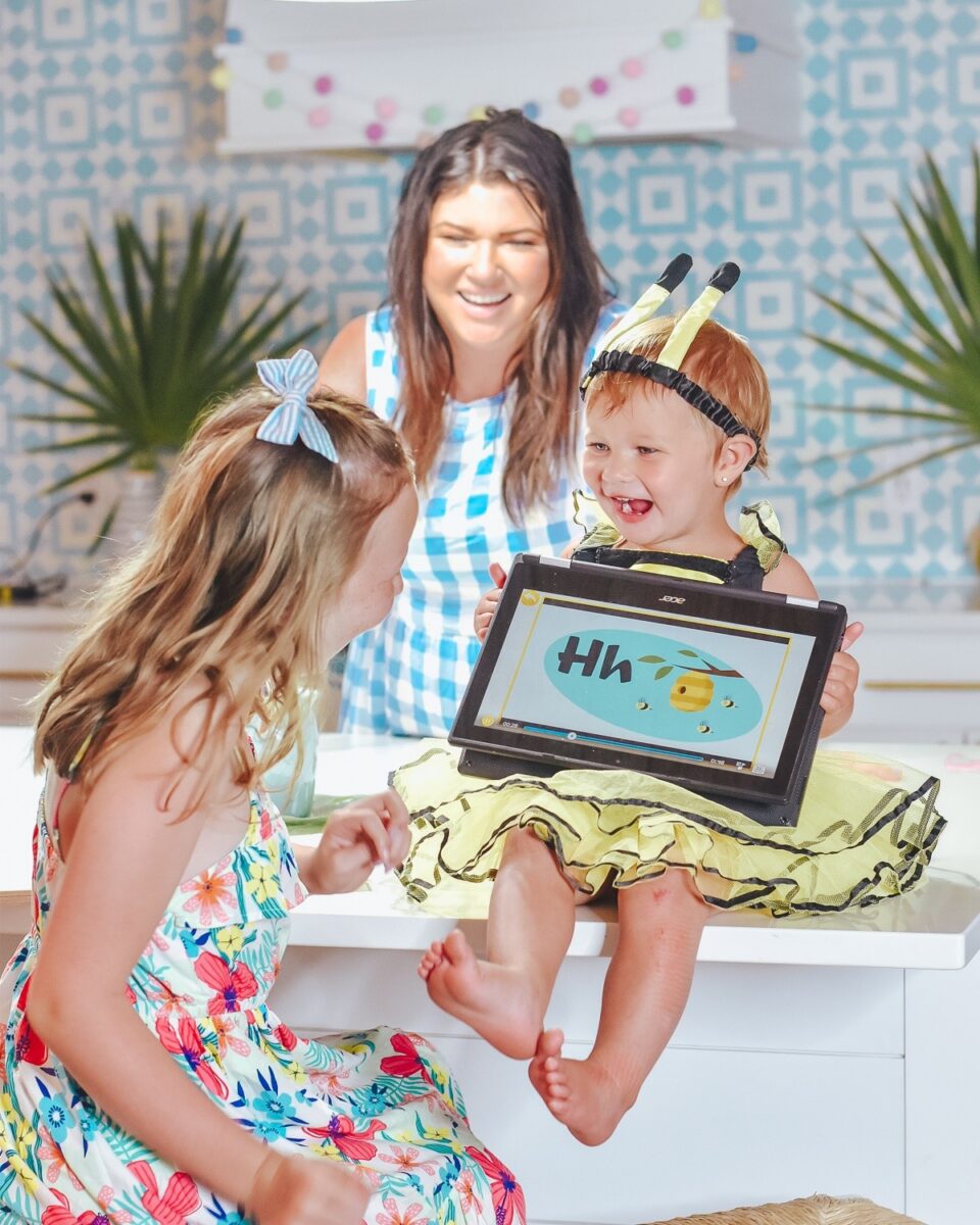 A smiling toddler in a bee costume sits on a counter, holding a tablet showing a 'Hi' slide while two other kids and an adult look on.