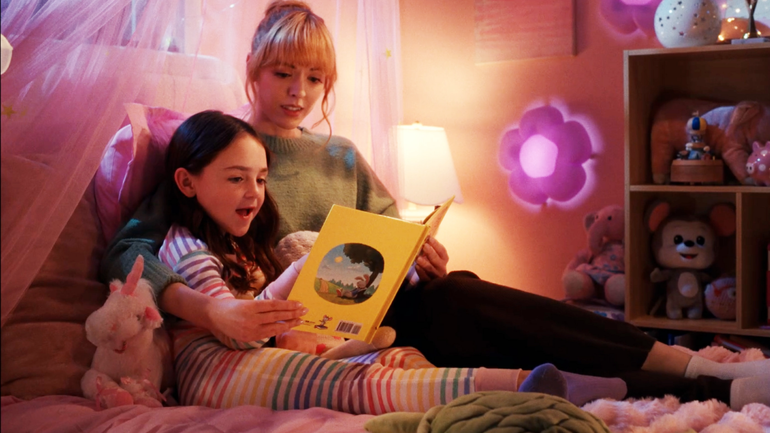 A woman and a young girl read a yellow children’s book together on a pink bed in a cozy, toy-filled bedroom.
