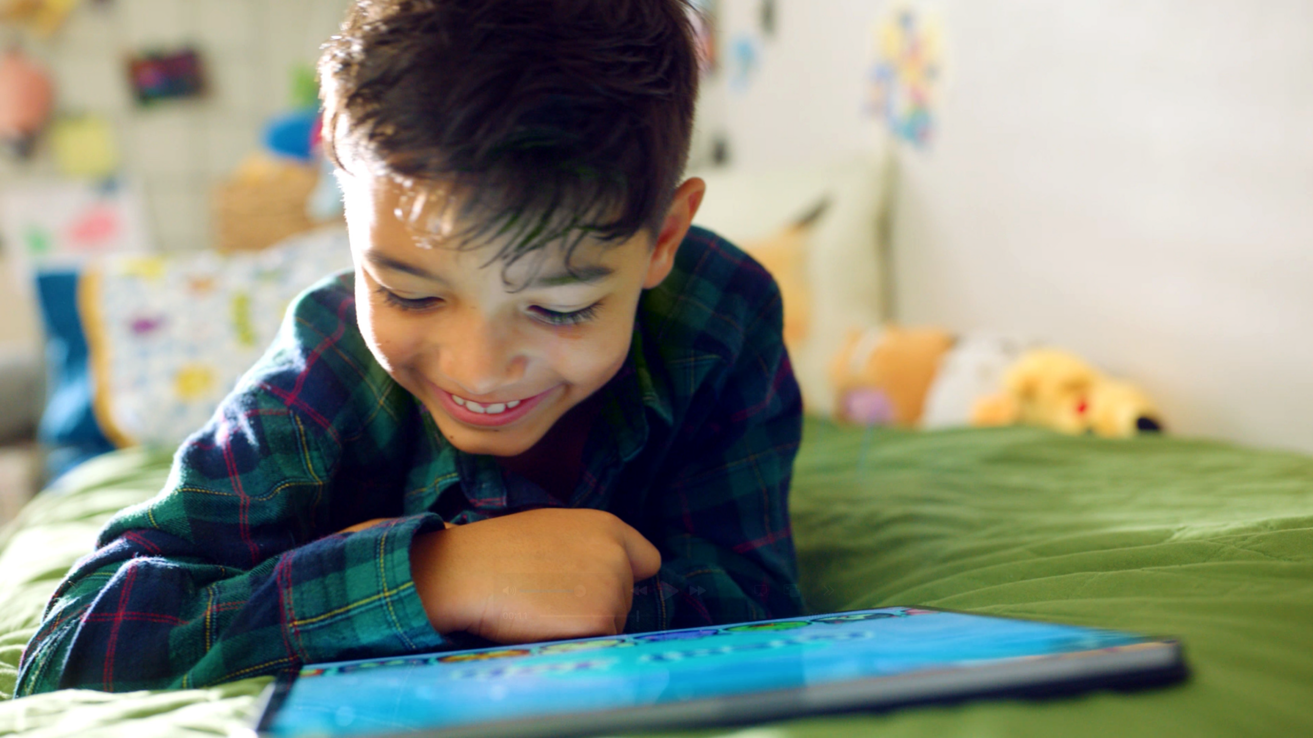 Smiling child in a green plaid shirt leaning over a tablet on a bed with a soft, colorful background.