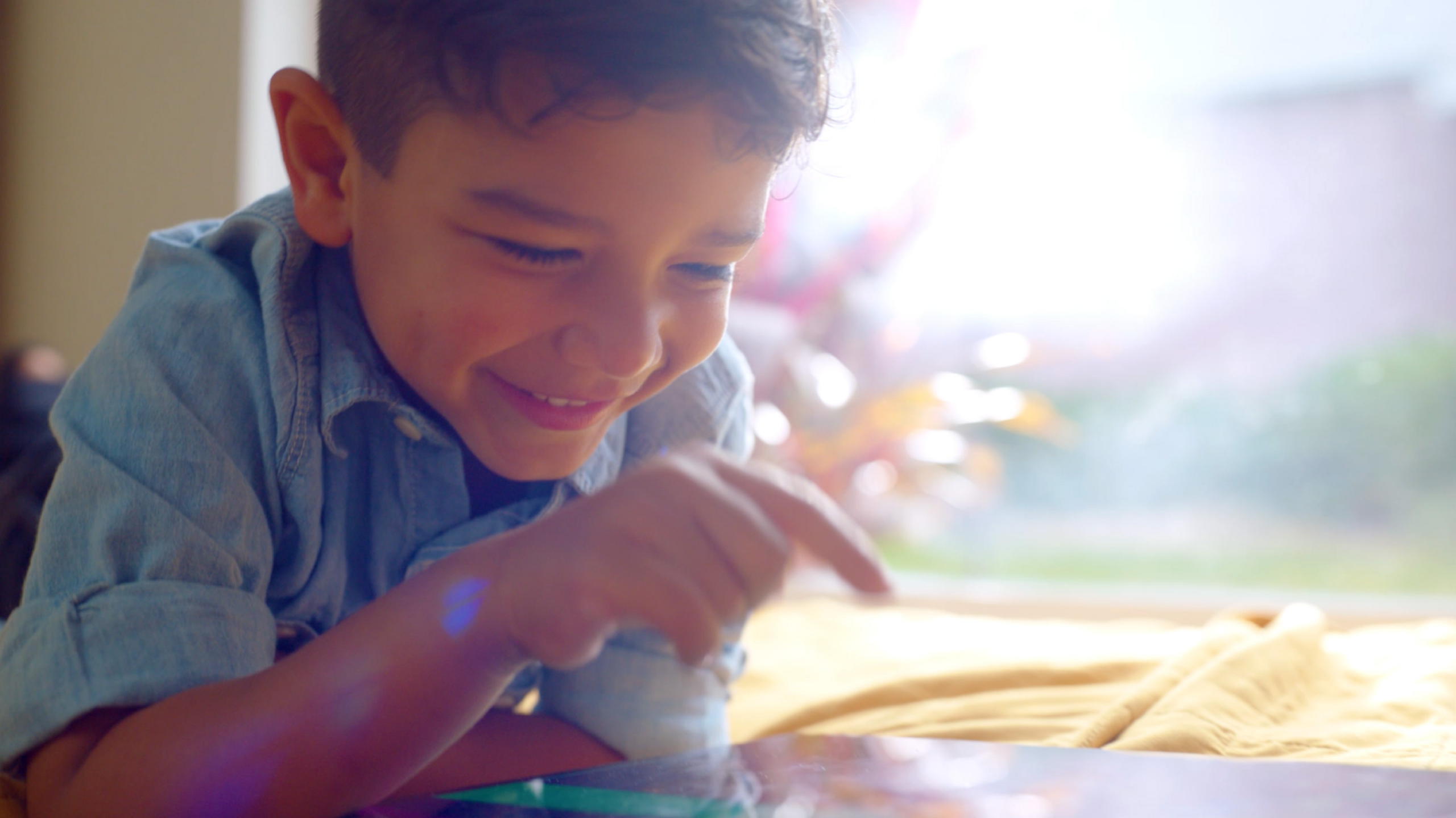 Close-up of a smiling boy in a denim shirt leaning over a table by a bright window with soft backlight.