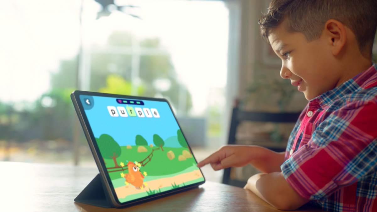 Boy in a plaid shirt plays a colorful tablet game with a cartoon landscape on screen.