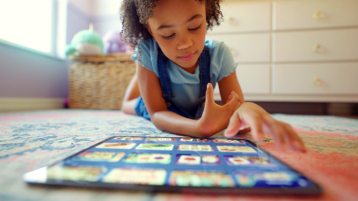 Child wearing blue shirt and denim overalls lies on a colorful rug, tapping a tablet screen with app icons visible.