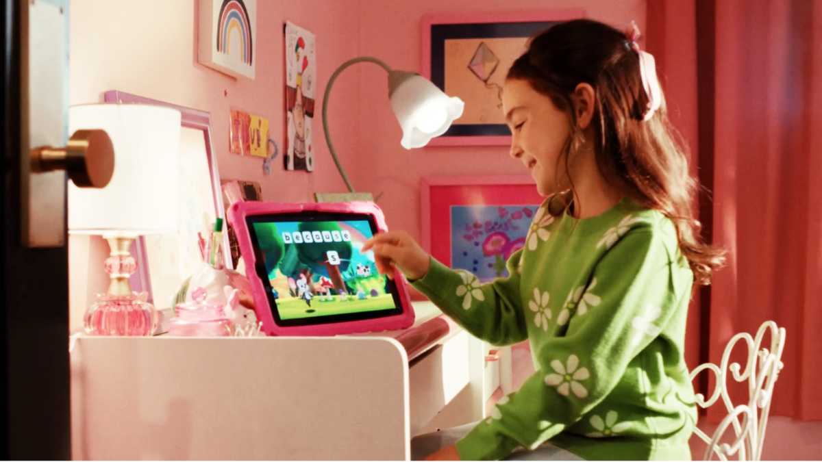 Girl in a pink-tinted room taps a pink tablet on a desk, engaged with the screen.