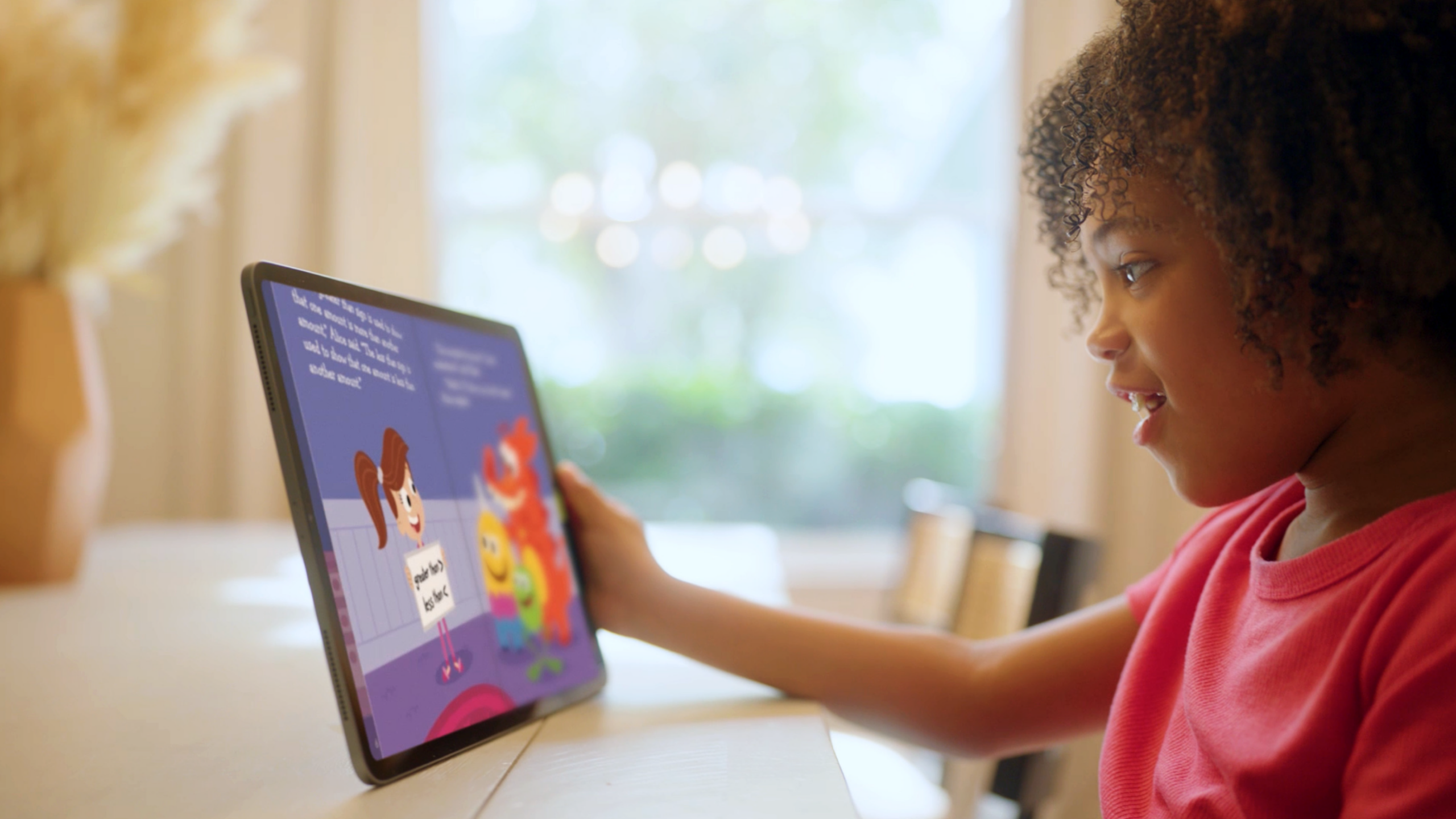 Child in a red shirt uses a tablet at a table, screen showing colorful cartoon characters.