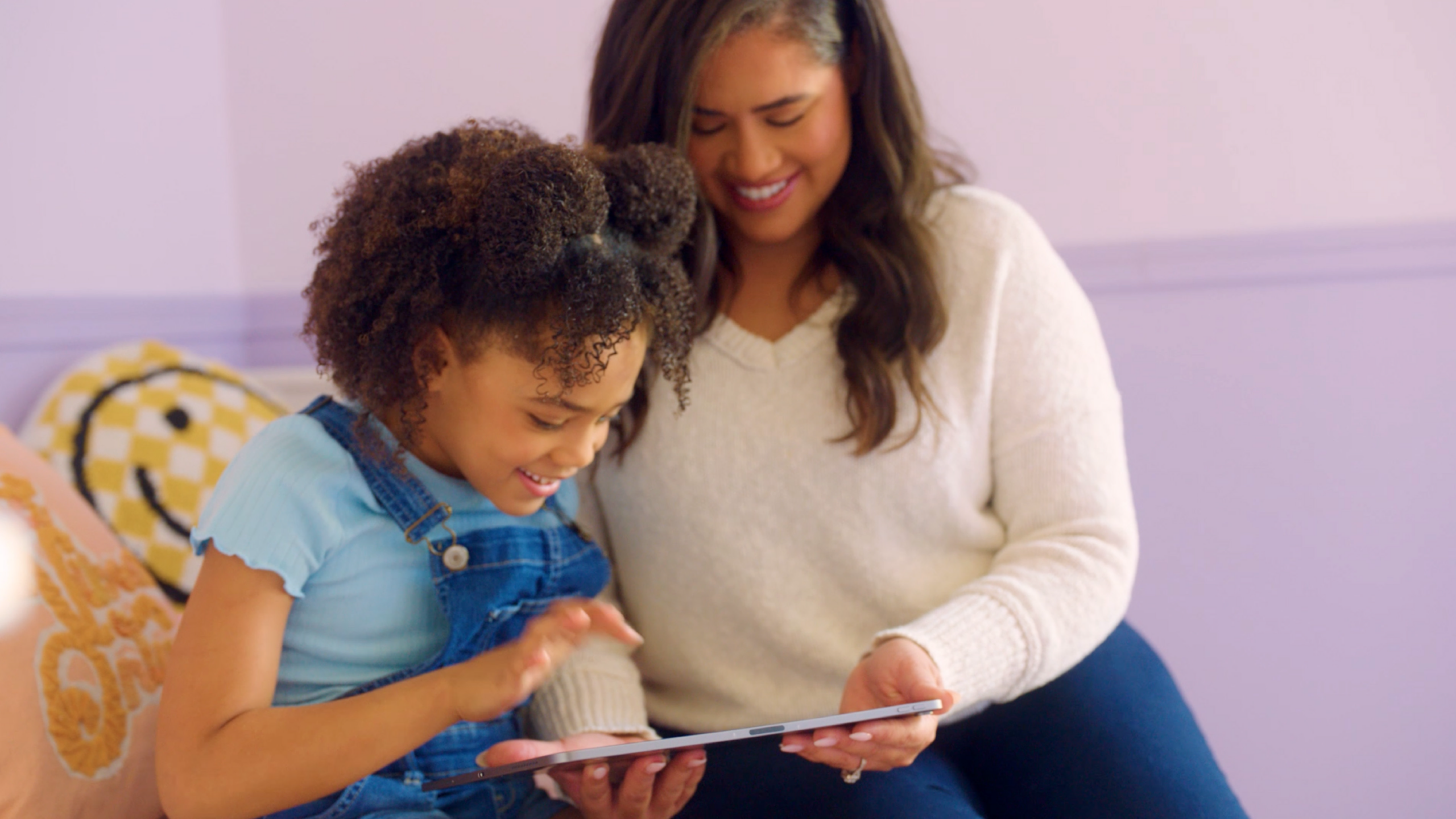 A young girl and an adult woman sit together looking at a tablet, smiling.