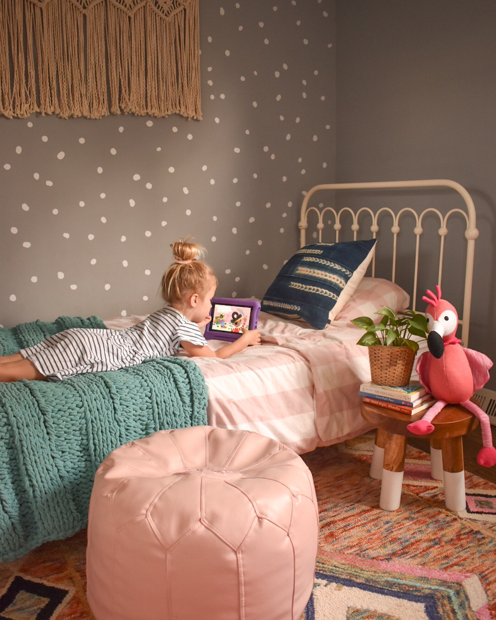 Child lying on a pink quilted bed using a tablet, with a teal knitted blanket and dotted wall behind her.