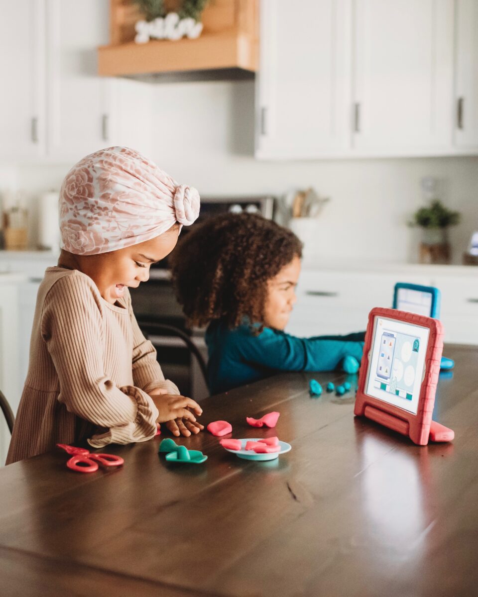 Two children sit at a wooden table making colorful clay shapes; one wears a pink patterned headscarf.