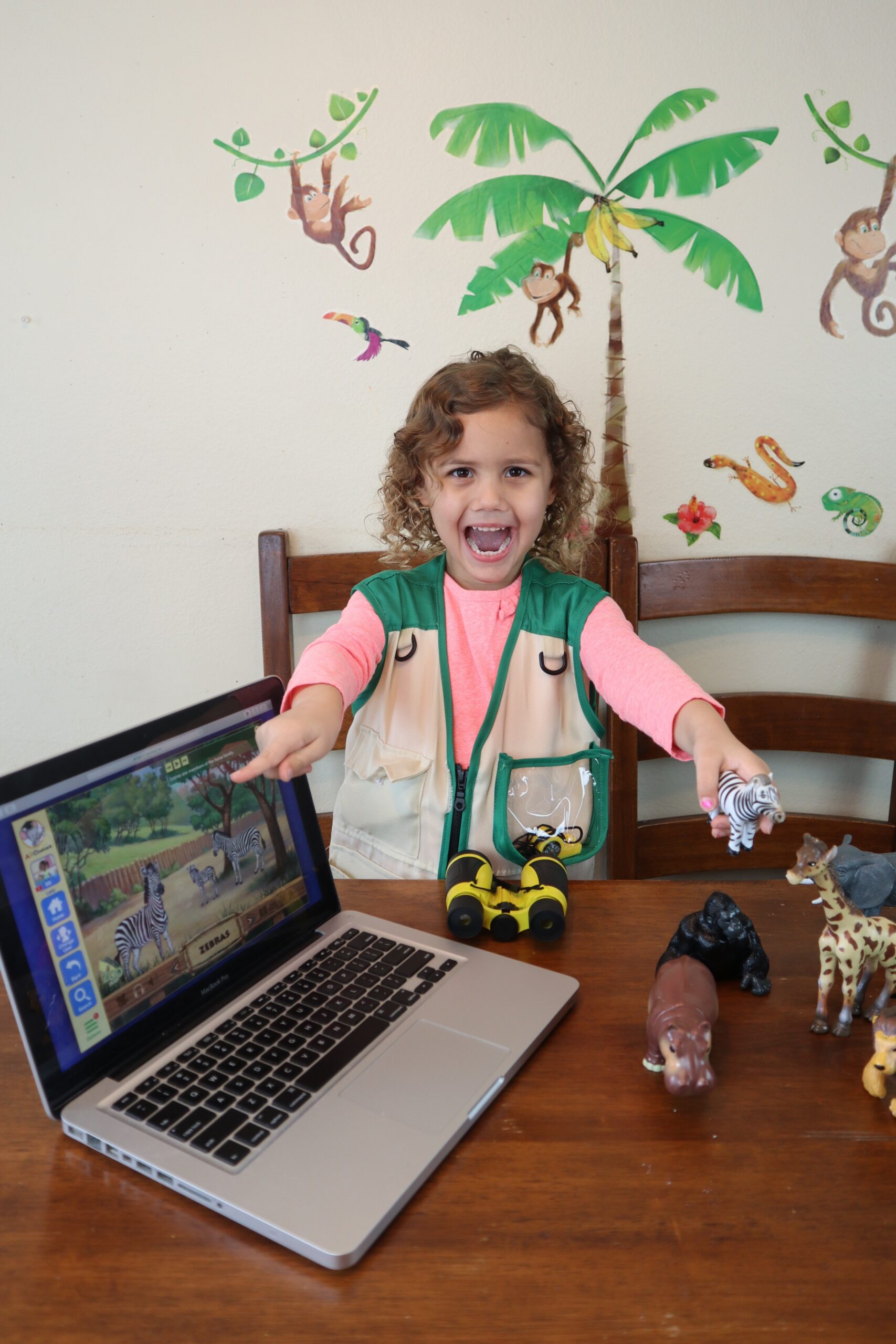 Smiling child with curly hair sits at a table with a laptop showing zebras, surrounded by jungle-themed toys and mural art.