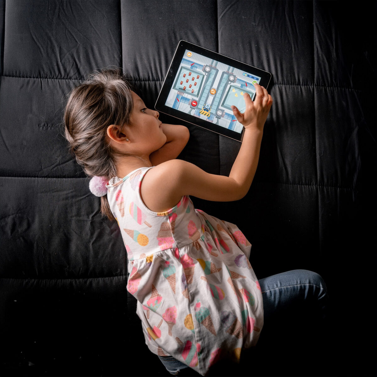 Girl lying on a dark couch, tapping a tablet showing a colorful game screen.Prefer concise alt text without stray tags.