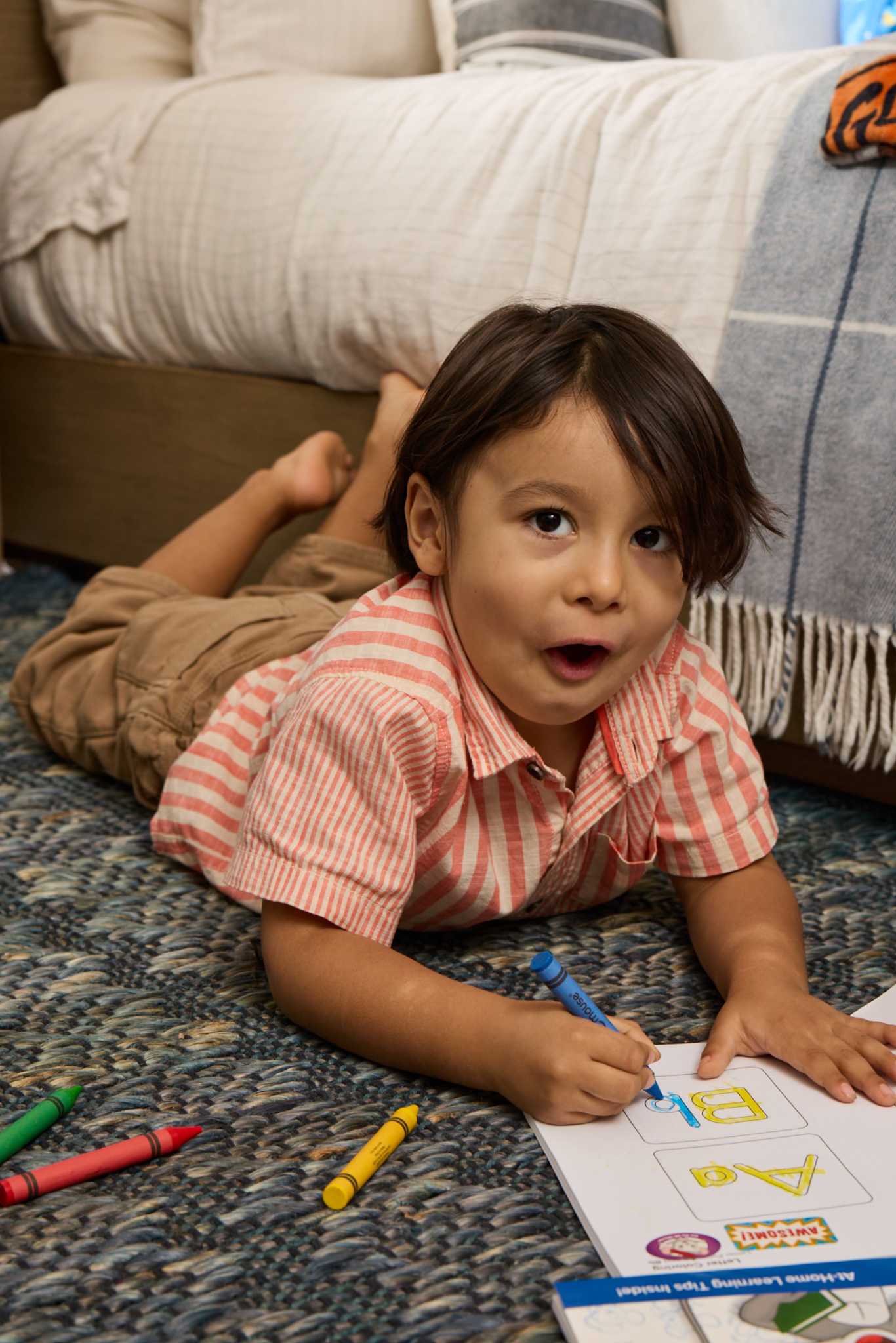 Young child lying on a patterned rug, drawing with a blue marker on a colorfully illustrated board beside scattered markers at home.