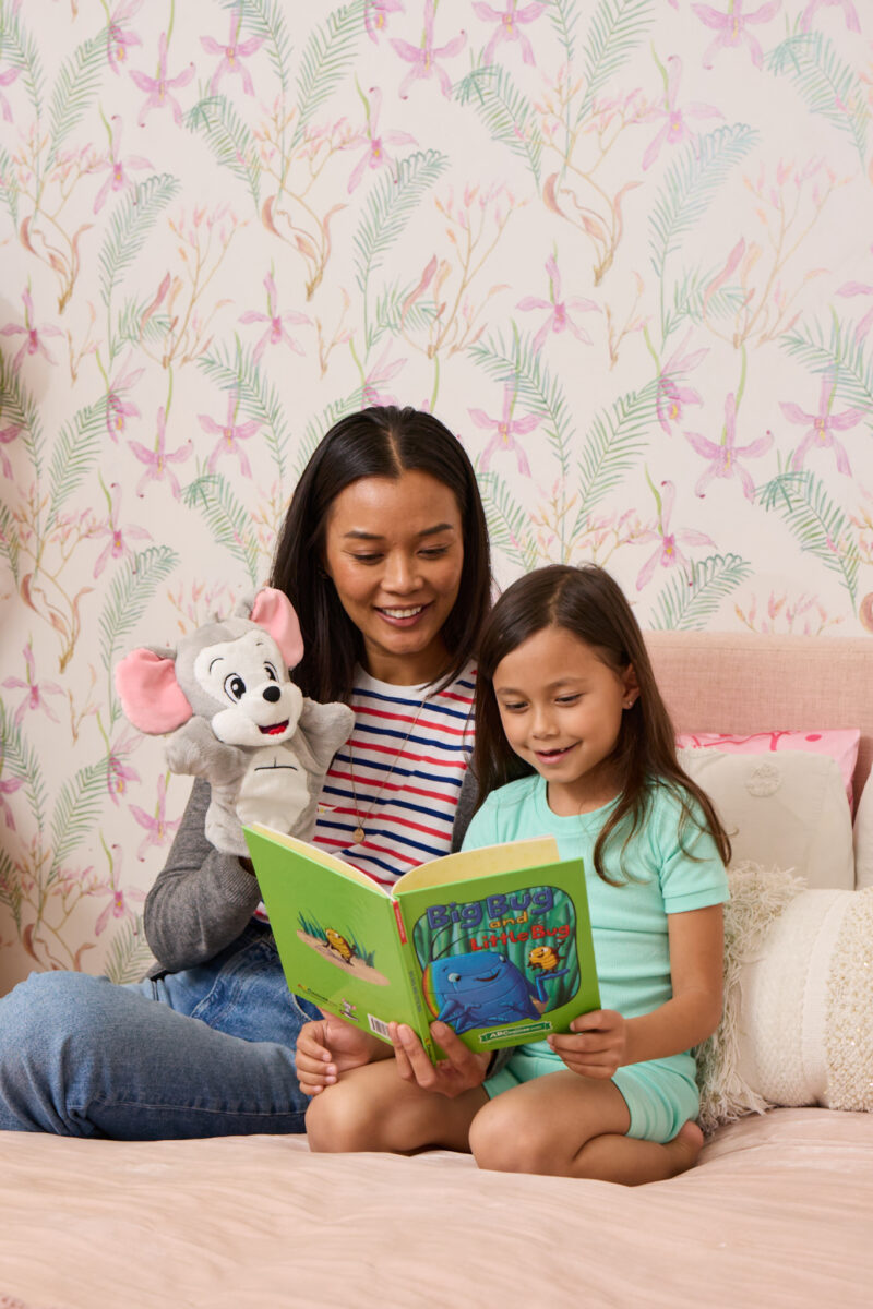 A woman and young girl sit on a bed reading a bright green children's book together, with a gray stuffed elephant nearby.