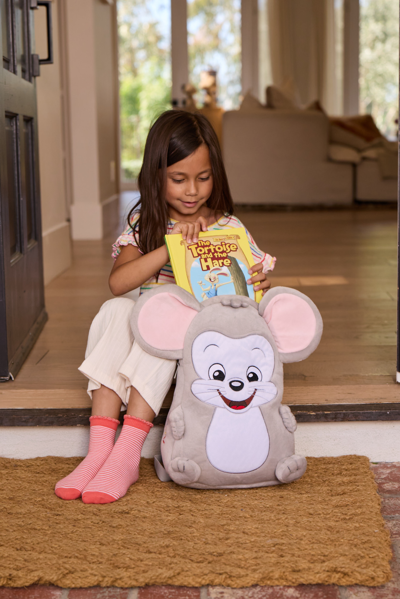 Young girl in a striped shirt sits on a doorstep reading “The Tortoise and the Hare” while hugging a large gray mouse plush.