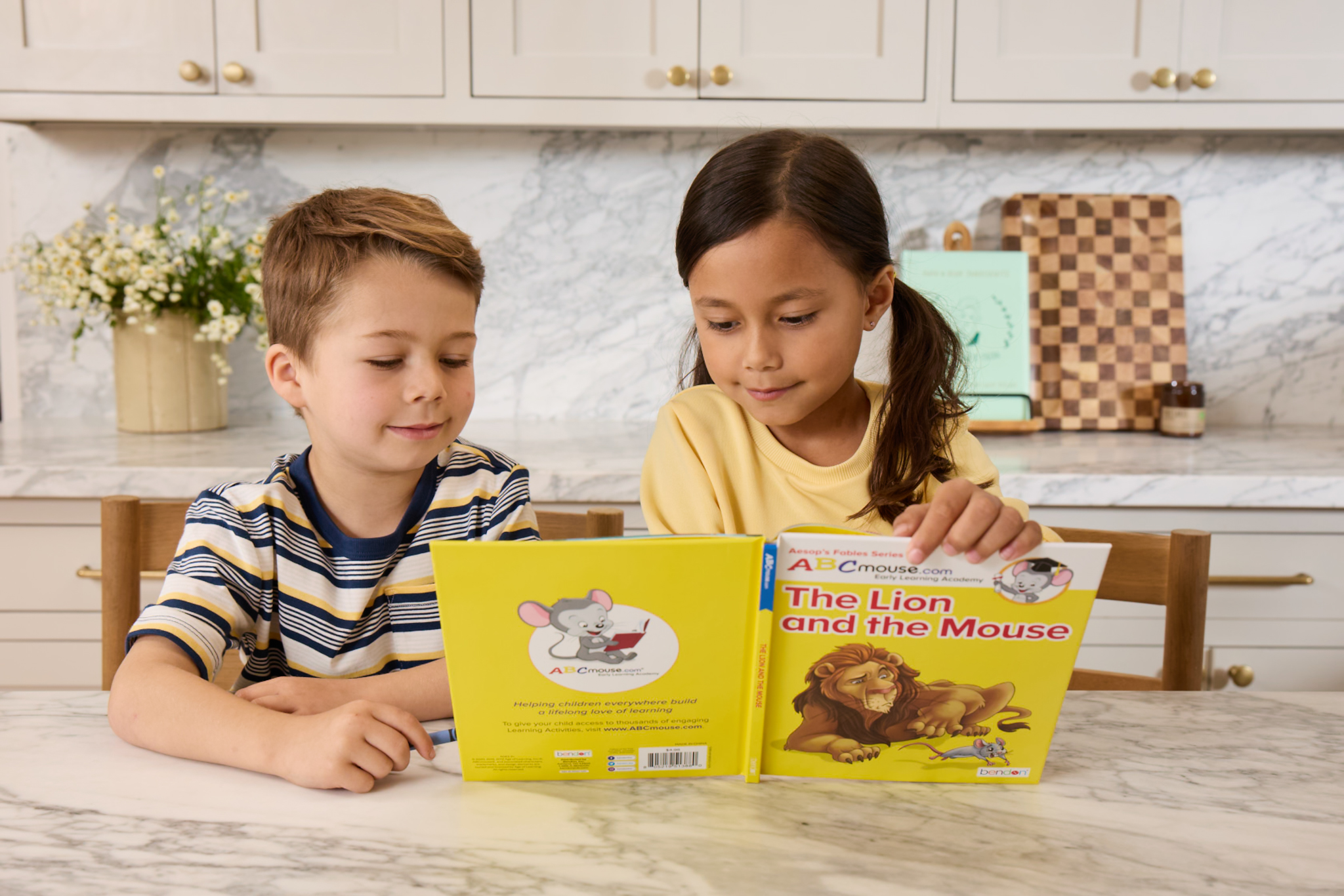 Two children sit at a marble kitchen table reading a bright yellow children's book together on a sunny day in a modern kitchen.