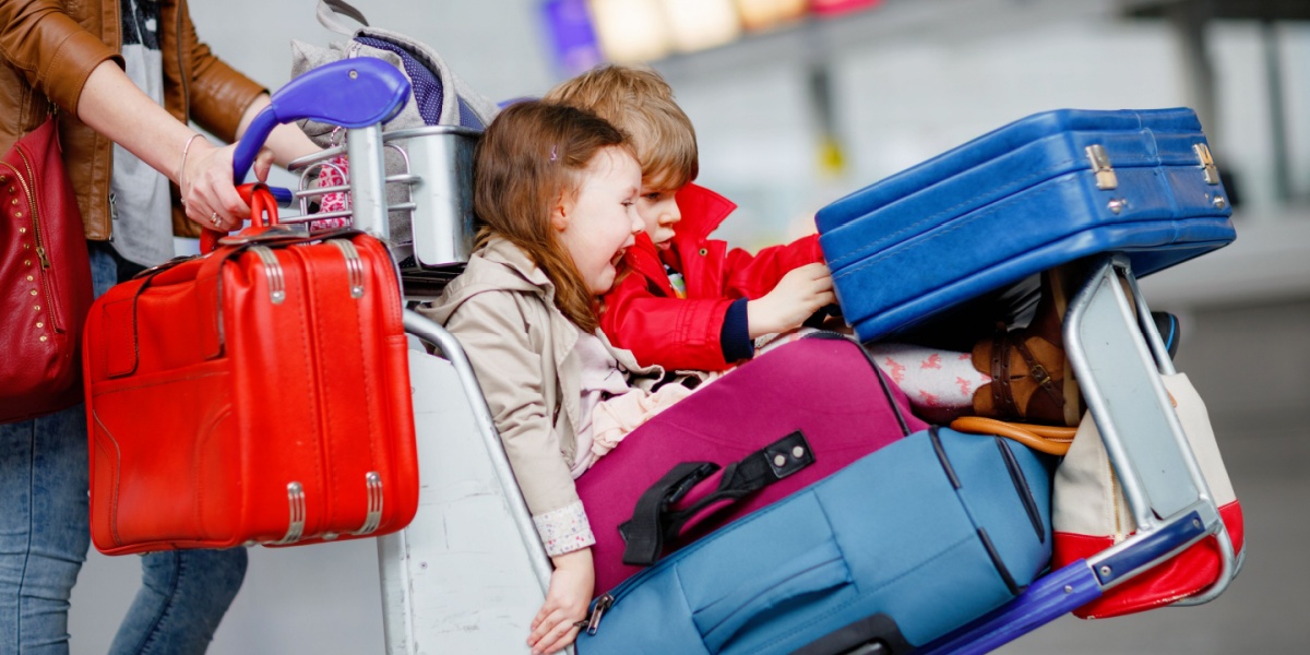 Two young children sit on an airport baggage cart surrounded by colorful suitcases while an adult pushes the cart through a terminal, suggesting family travel or a vacation journey.