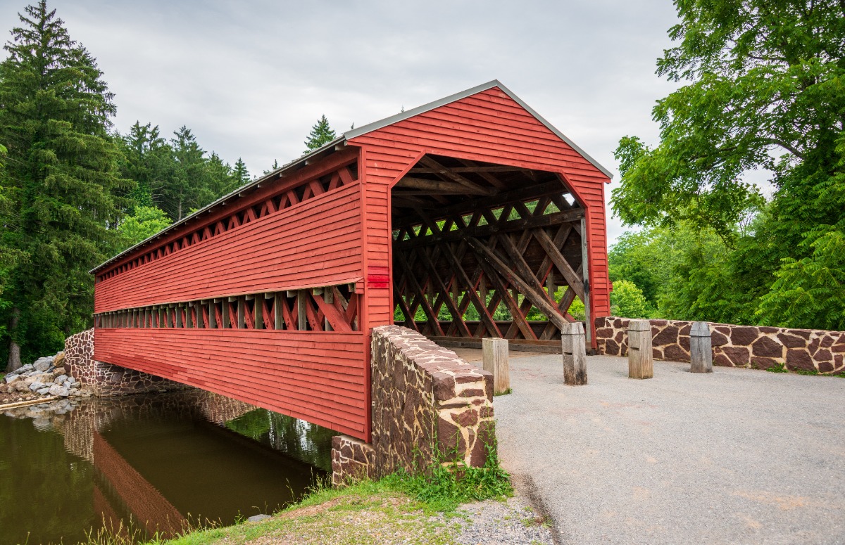 Red wooden covered bridge spanning a calm river, surrounded by green trees and a cloudy sky, with stone abutments nearby.