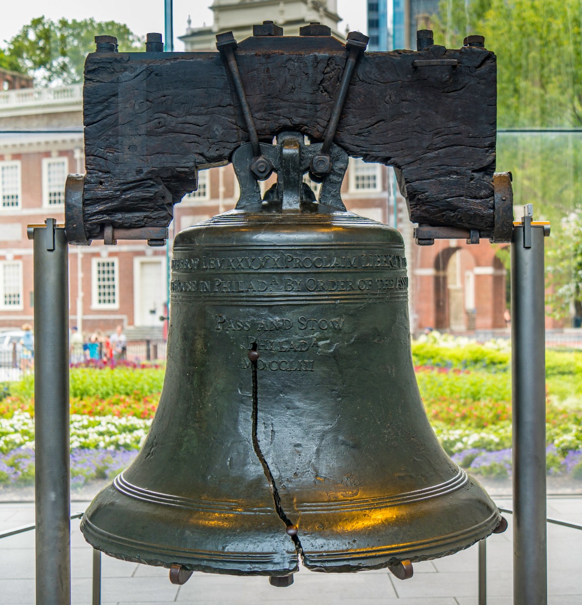 The cracked Liberty Bell hanging from a dark wooden yoke in front of a glass wall and brick buildings, in a museum setting.