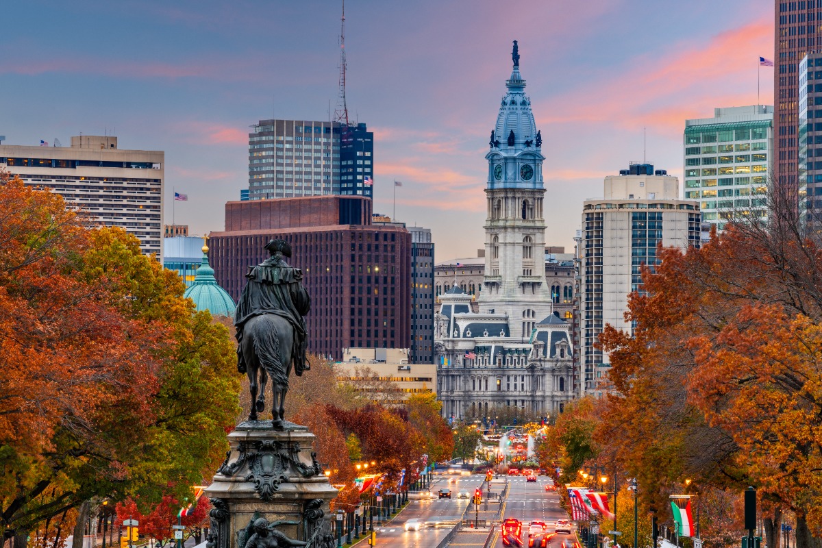 Equestrian statue on a carved pedestal along a tree-lined city street in autumn, with Philadelphia City Hall in the distance.