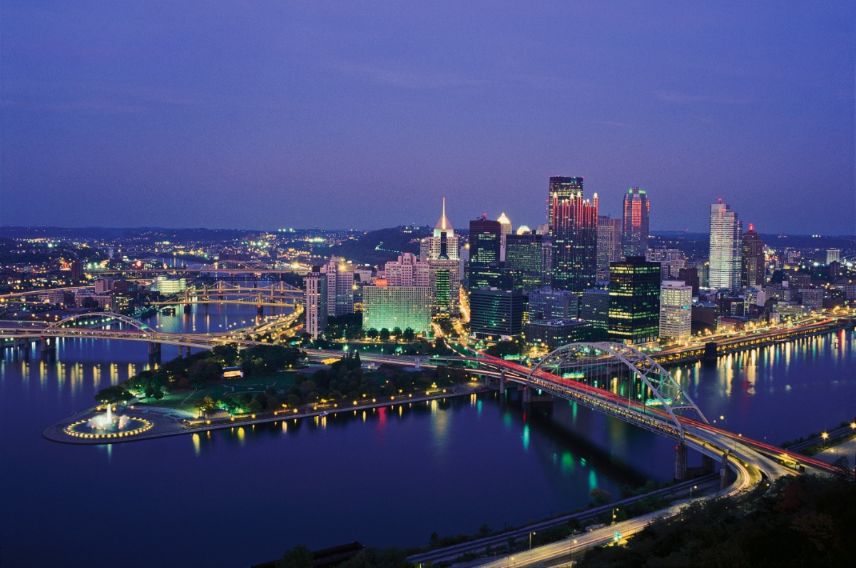 Evening city skyline over a calm river with illuminated bridges and tall buildings.