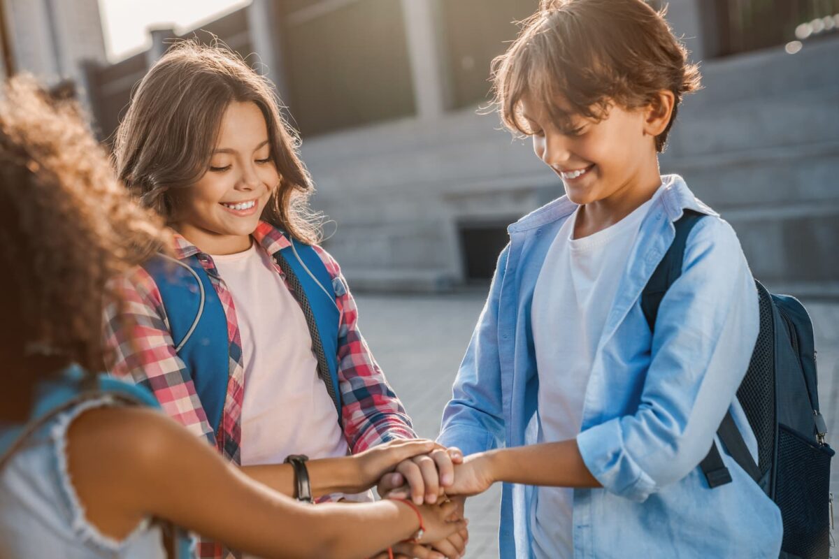 Three kids with backpacks join hands in a show of friendship outdoors, smiling at each other during a school moment.