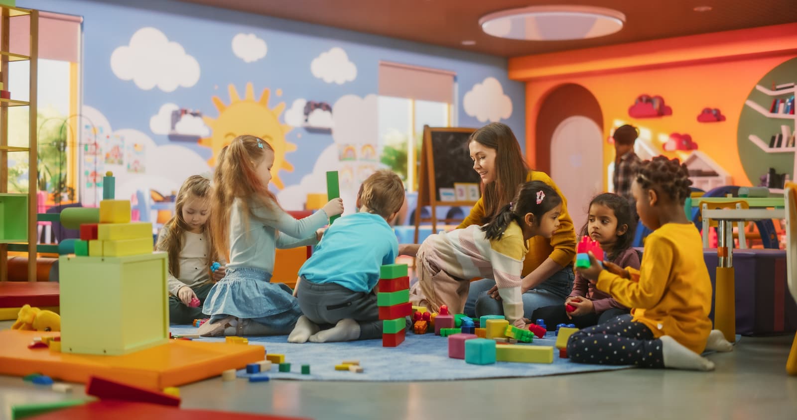 Children building a tower with large foam blocks on a blue rug in a colorful playroom as a caregiver watches.