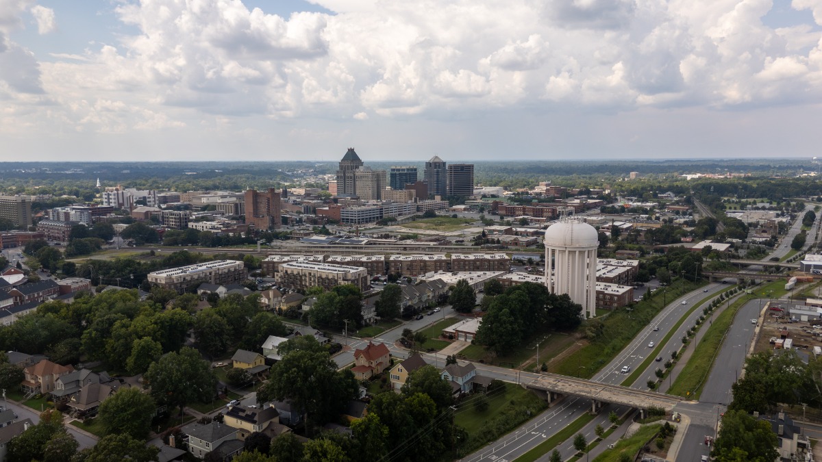 Aerial view of a city with a white water tower, highway overpass, and a mix of low-rise and mid-rise buildings under a cloudy sky, taken from above.