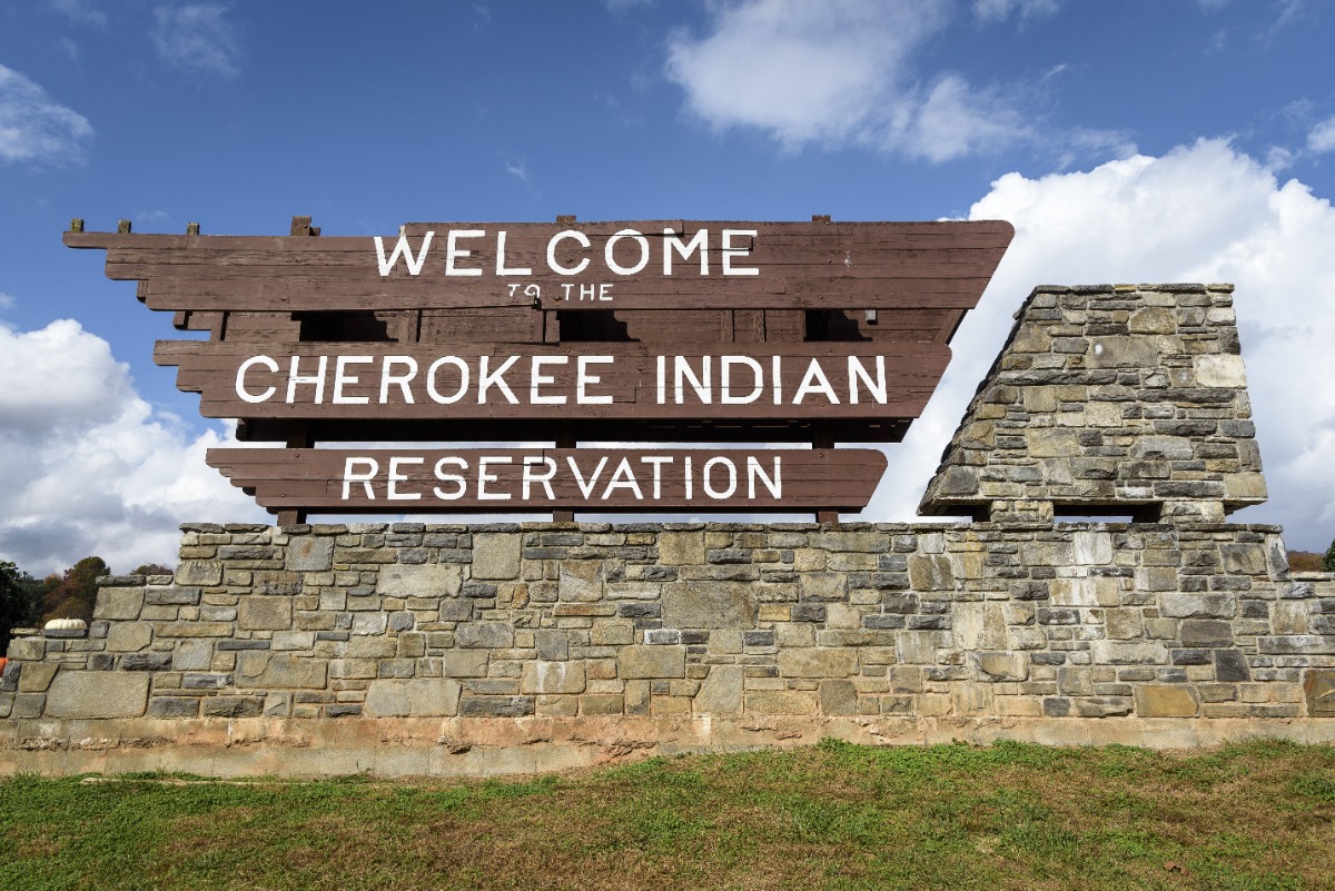 Brown wooden sign reading 'Welcome to the Cherokee Indian Reservation' mounted on a stone wall under a blue sky with clouds