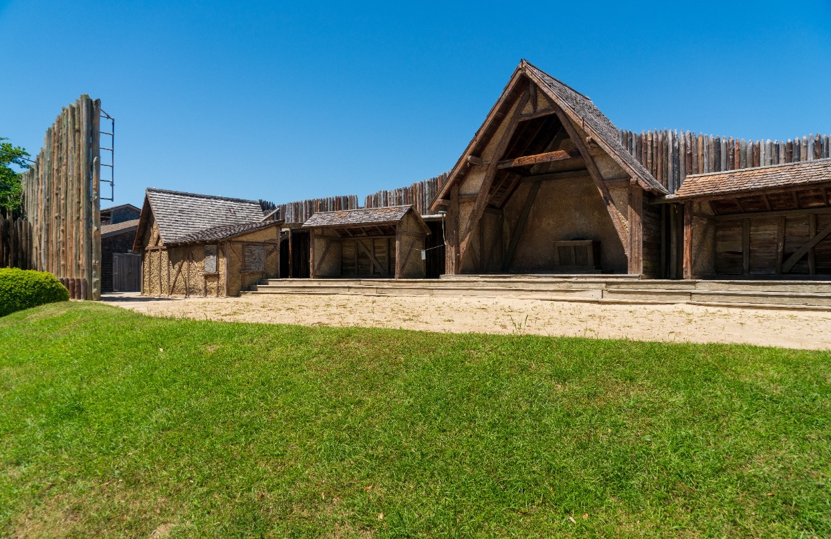 Reconstructed wooden fort with a palisade wall, thatched huts, and a central open stage on a sunny day