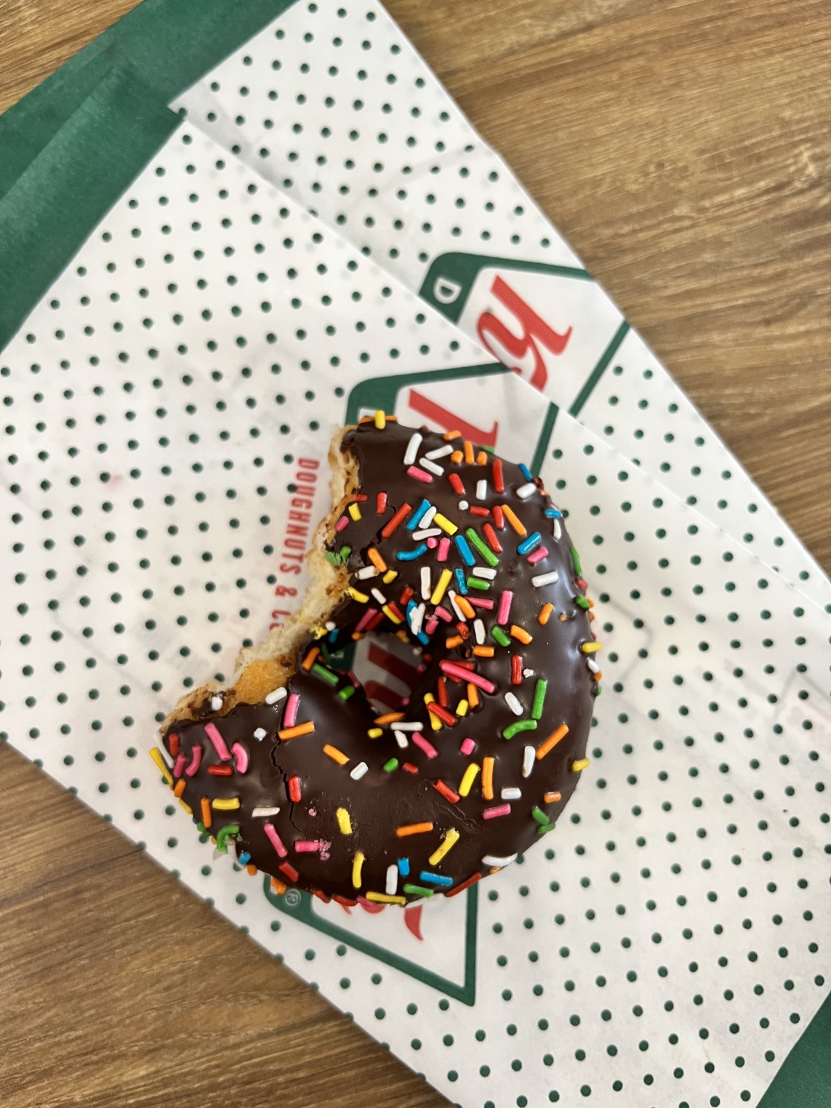 Chocolate donut with rainbow sprinkles on a white perforated bakery wrapper on a wooden table.