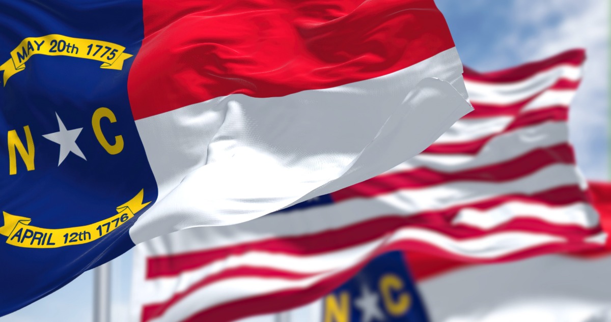 North Carolina state flag in the foreground with the American flag waving in the background outdoors; NC seal and dates visible on the blue field with a white star and letters N and C.